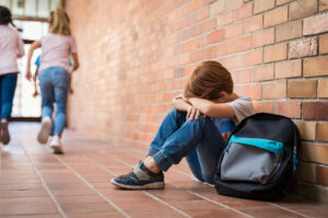 A young boy sitting alone against a brick wall at school. Representing how child anxiety can manifest in different environments. Start working with a child therapist in Katy, TX today!