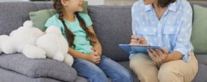 A child therapist & young girl sitting on a gray couch talking. To learn more about co-parenting after divorce, get started with our child counseling in Katy, TX. We are here to support you. 