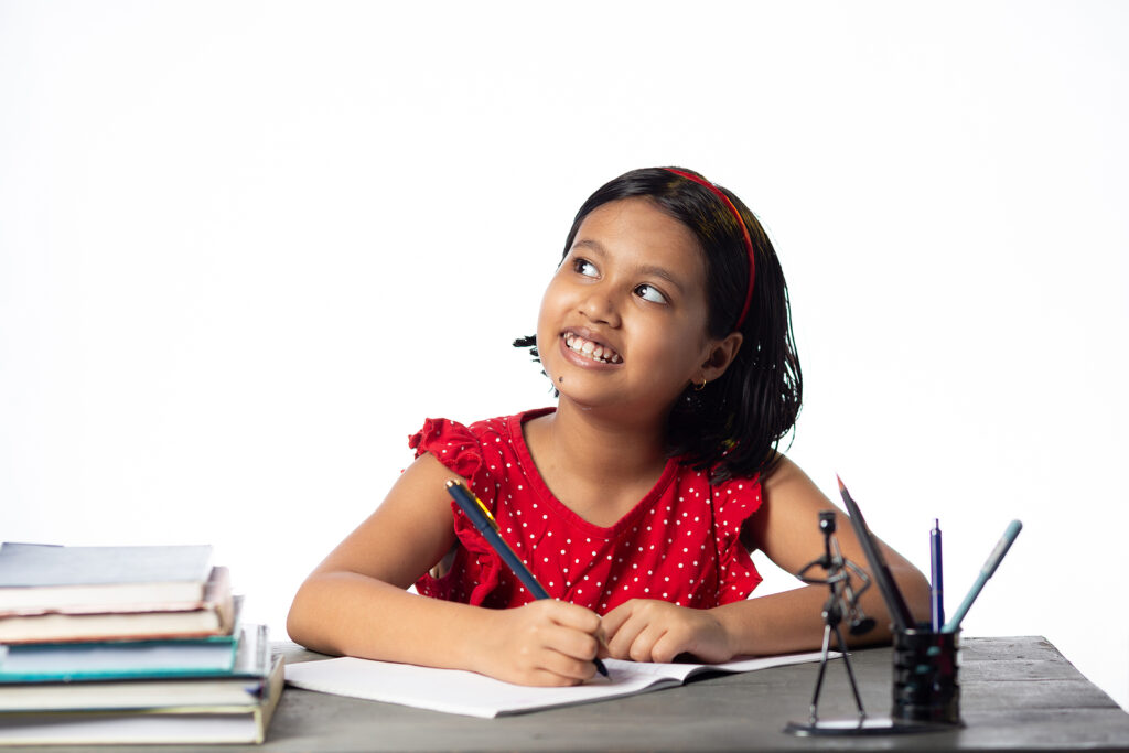 A young girl looking up, smiling & doing her homework. If your child struggles with school anxiety, reach out to begin child anxiety counseling in Katy, TX today. We can help!