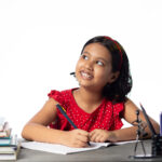 A young girl looking up, smiling & doing her homework. If your child struggles with school anxiety, reach out to begin child anxiety counseling in Katy, TX today. We can help!