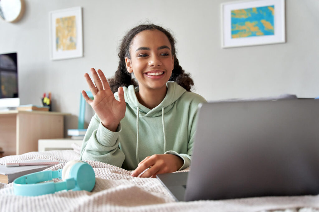 A teen girl waving at her laptop & smiling. Representing how online therapy for teens in Katy, TX can help your child with at-risk behaviors. Call today to begin!