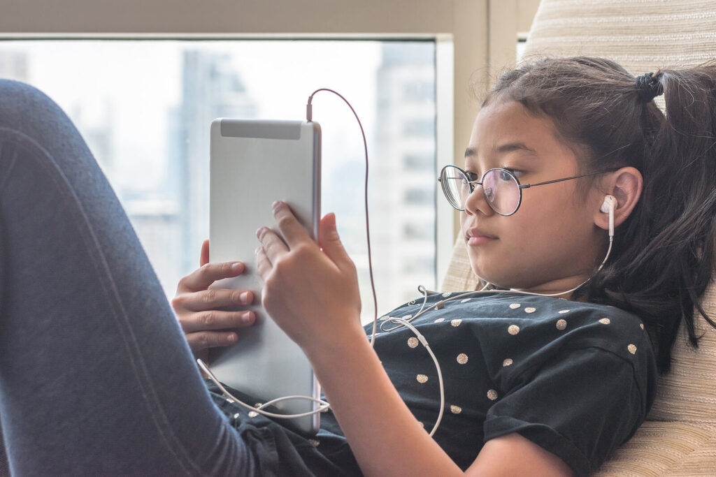 A young girl resting while watching an iPad and listening to headphones in her ears. Representing how the overuse of technology can increase anxiety in children. Reach out today to get started with out child counseling for anxiety in Katy, TX. 