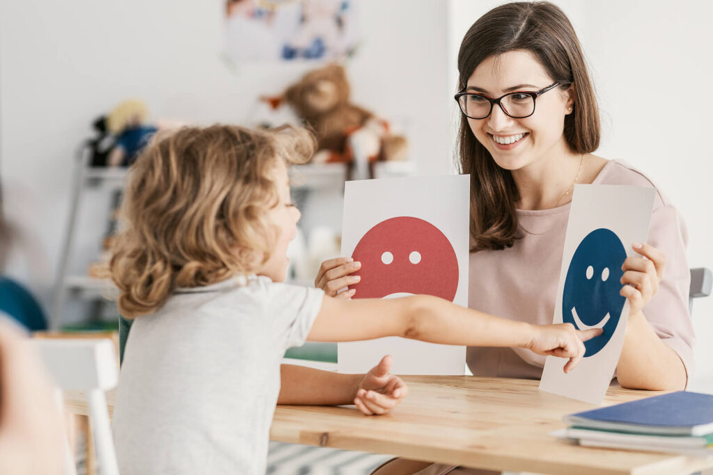 A child therapist holding two emotions on paper & a child pointing to the happy face. Want to learn more about behavioral therapy for children in Katy, TX? Reach out to us today!