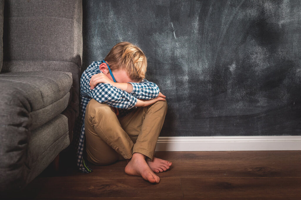 A young blonde boy sitting in the corner while hiding his face in his arms. Child behavioral therapy in Katy, TX can help your child build emotional resiliency. Call today to get started. 