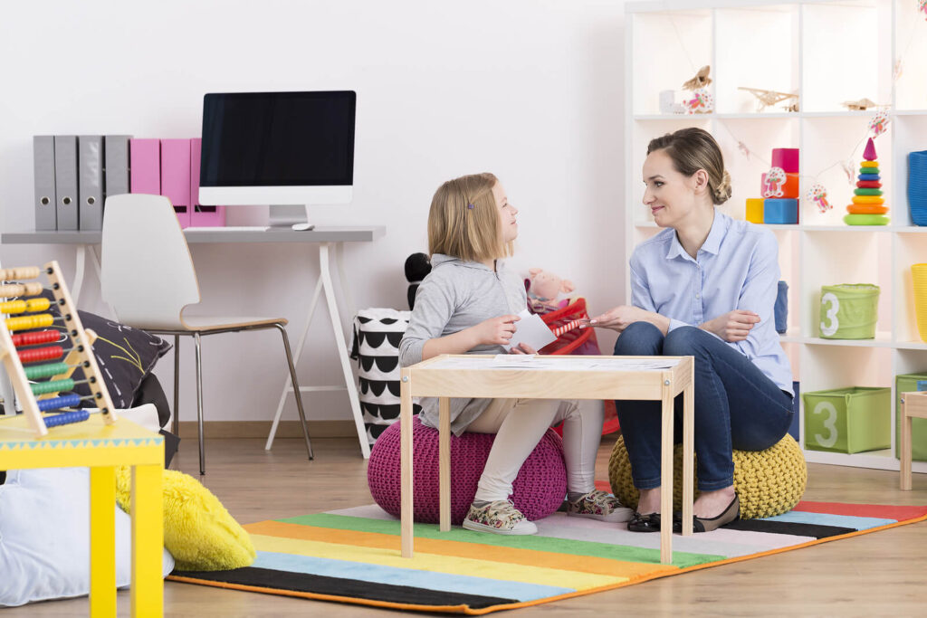 A child therapist sitting with a young girl at a play table during a session. If your child struggles with big emotions, get started with a child behavioral therapist in Katy, TX today. We can help your child build resiliency. 