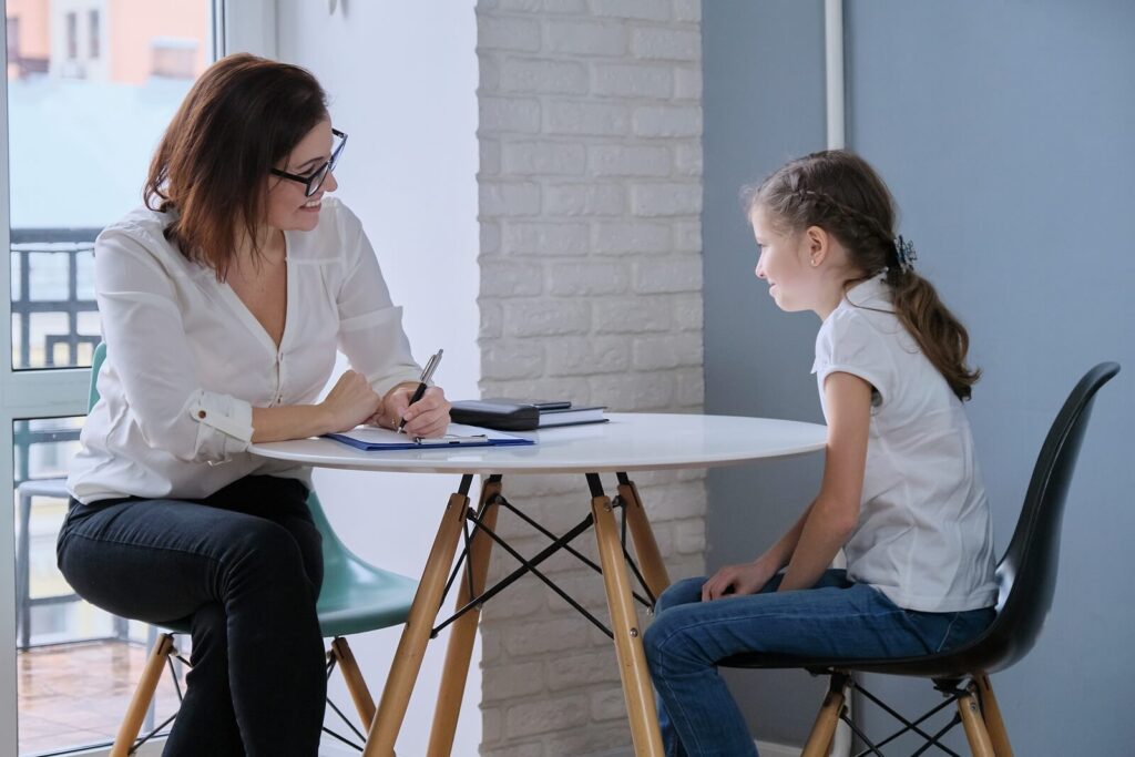 A child sitting at a white table with a child therapist who is taking notes. Representing how child therapy in Katy, TX can help your family as a whole. Reach out to our therapists today. 