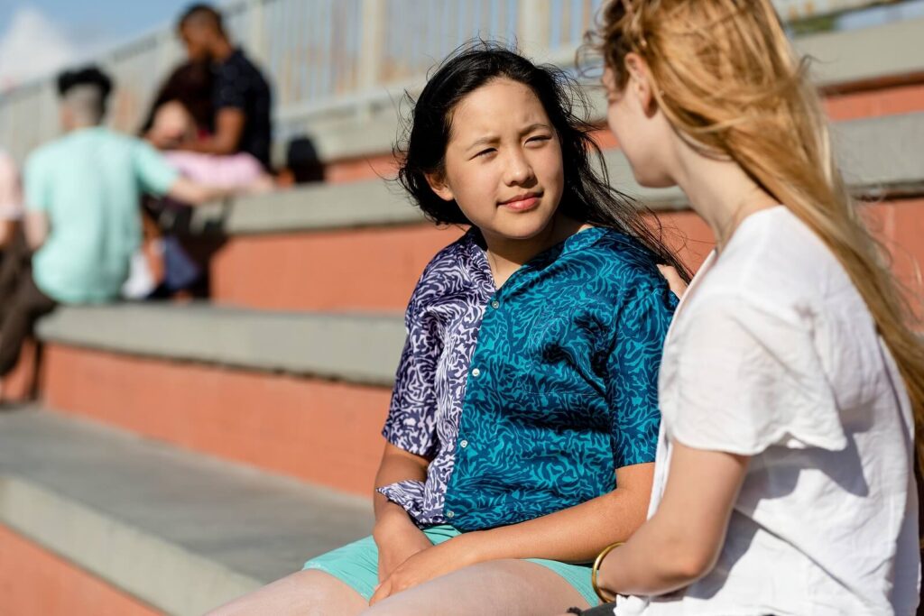 A young teen girl wearing a colorful shirt sitting on the bleachers with a friend. If you suspect your teen is participating in at-risk behaviors, a teen therapist can help. Call us now to begin!