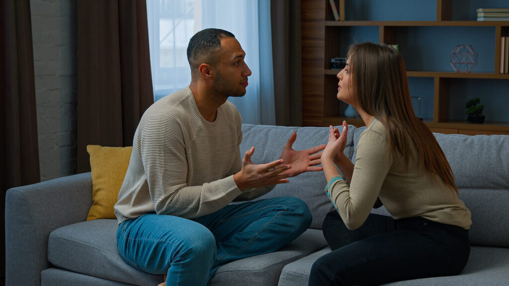 A couple sitting on a blue couch while having a tense conversation. Couples counseling in Katy, TX helps relationships thrive again. Read our blog here for more information on encouraging willingness to start therapy!