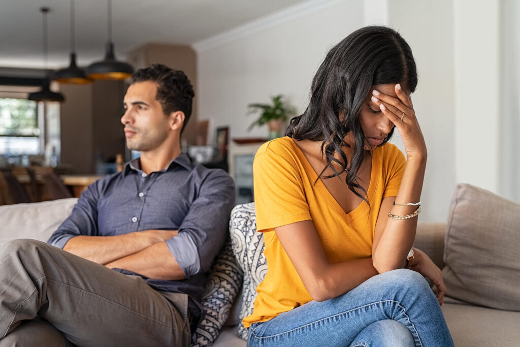 A couple sitting away from each other on the couple while the woman hides her face with her hands. A couples therapist in Katy, TX can help you improve your communication! Reach out today to get started.