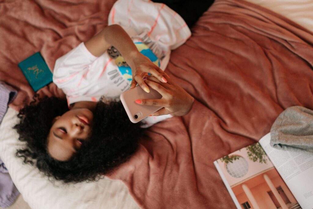 A young Black teen laying on her bed while scrolling on the phone. Representing how social media comparison is harmful to teen's mental health. Discover how teen therapy in Katy, TX can help your teen find balance. 
