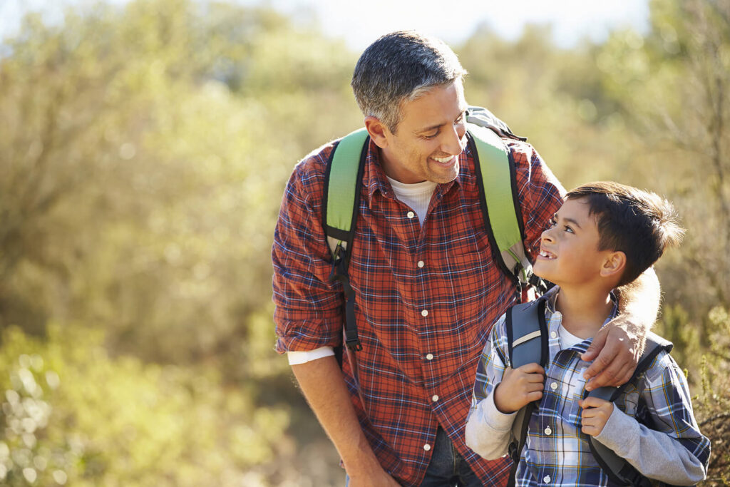 A father and son both wearing backpacks & smiling at each other during a hike. Child therapy in Katy, TX can help with grief after the floodings. Get personalized support for your child today. 