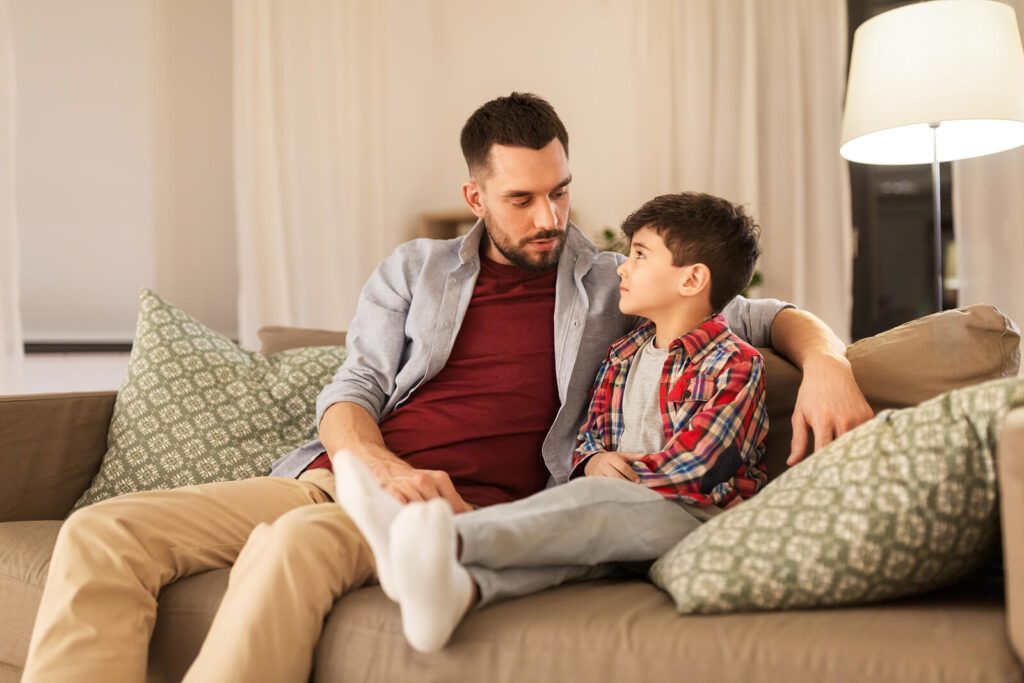 A father sitting on the couch talking to his young son. Child counseling in Katy, TX can help your whole family after a traumatic event. Help your child find peace today.