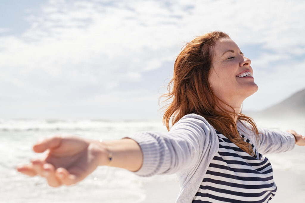 A redhead woman by the beach throwing her arms out in relief. Looking for LENS therapy near me? Discover how our LENS neurofeedback in Katy, TX can help your nervous system.