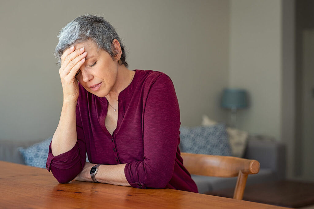 An older woman rubbing her forehead in distress sitting at a table. If you want to balance your nervous system, LENS therapy in Katy, TX can help. Learn more here!