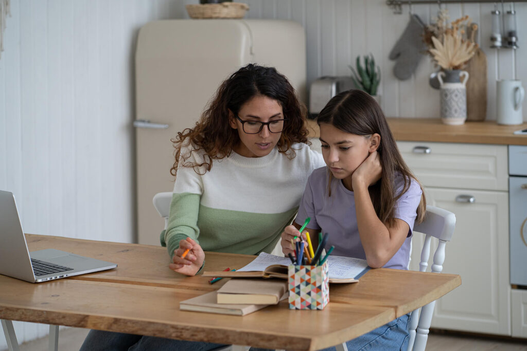 A mother sitting down with her daughter at the kitchen table doing schoolwork. Women often juggle the invisible load at home. Discover how LENS neurofeedback in Katy, TX can help!