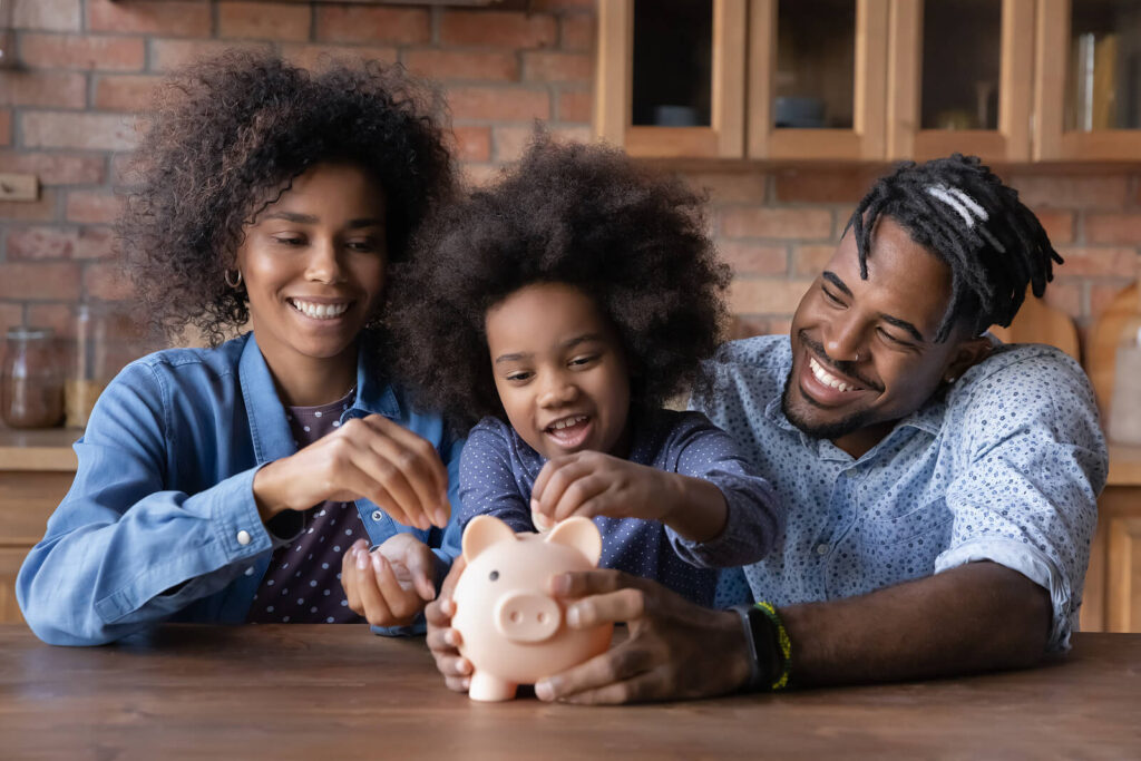 A happy Black family putting coins into a piggy bank on the table. Representing how WHCC's child therapy in Katy, TX is offered at more affordable rates. Get started today. 