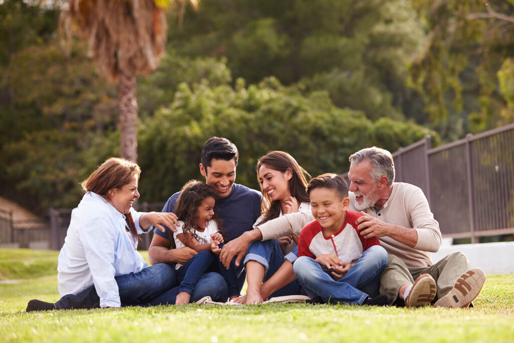 A happy Hispanic family all laughing together outside sitting in the grass. Our child therapists in Katy, TX would love to help your child thrive. Reach out today to get started. 