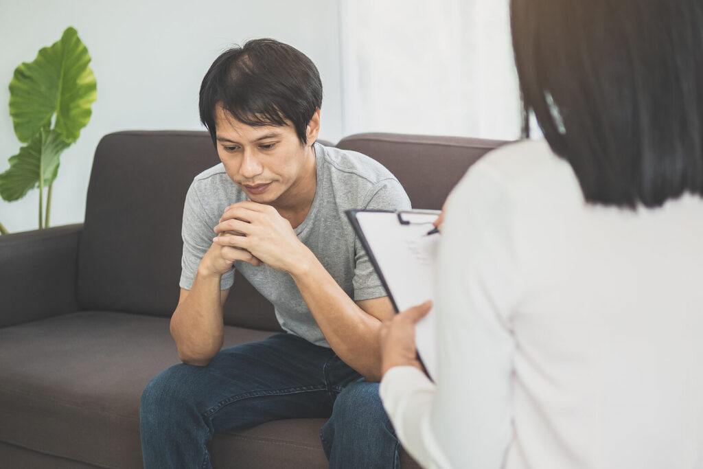 An Asian american Man holding his hands together while sitting on a gray couch. Finding an affordable therapist in Katy, TX can help you with stress, anxiety, relationship struggles & more. Reach out today to get started. 