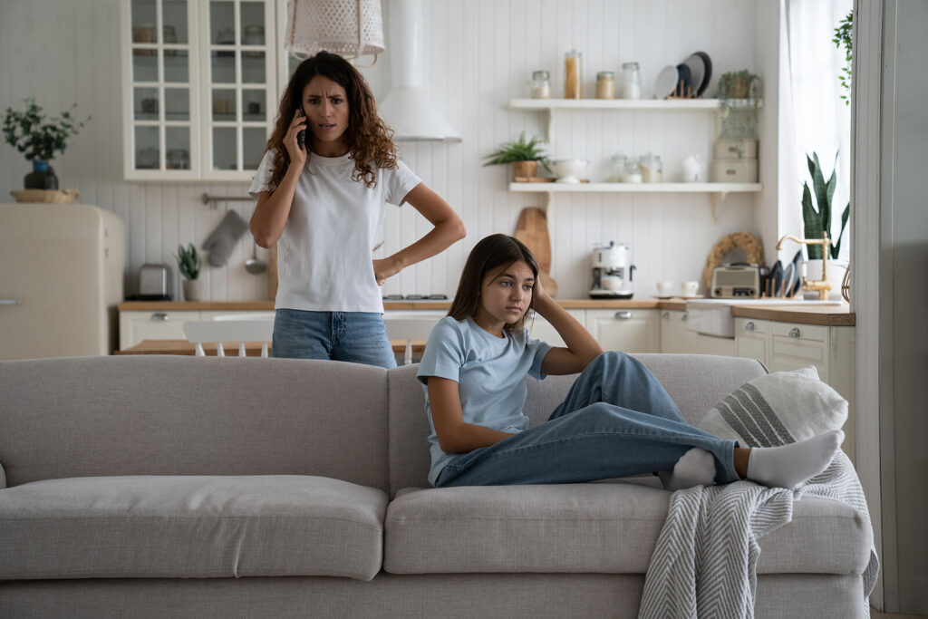 A mother standing talking on the phone while her daughter sits away from her on the couch. Representing how risky teen behavior can cause disruption in the family. Reach out today to begin teen counseling in Katy, TX.