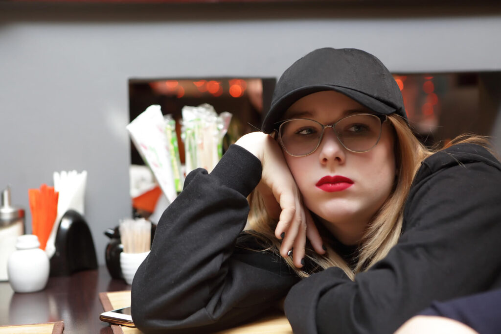 An annoyed teen girl wearing all black & red lipstick sitting at a booth. Teen therapy in Katy, TX can help your teen with at-risk behaviors. Learn more here!