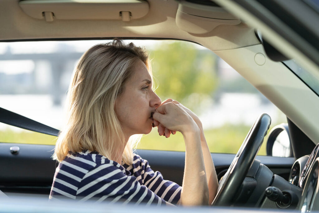 A woman sitting alone in her car while biting her nails. Representing how anxiety can impact school drop off, work & more. Get started with adult therapy in Katy, TX today.