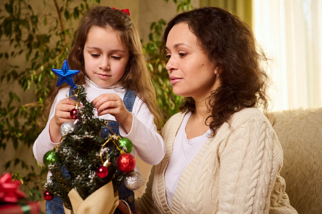 A Hispanic mother sitting next to her daughter decorating a Christmas tree. If you're struggling with holiday stress from managing it all, we're here to help. Get started with an adult therapist in Katy today.