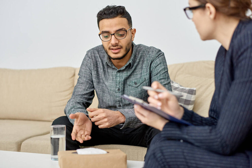 A Biracial man sitting on a cream colored couch talking to his therapist. A men's therapist in Katy, TX can help you rebalance before the new year. Call us today!