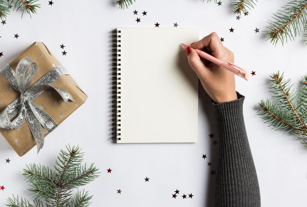 Individual writing in a blank notebook with a pink pen by pine needles & a present. An adult therapist in Katy, TX is here to support you in the new year. Discover more here!