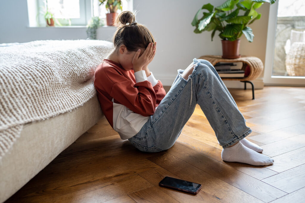 A teen sitting on the ground against a bed hiding their face with their hands. Social media usage can affect your teens mental health. Counseling for teens in Katy, TX can help them thrive. 