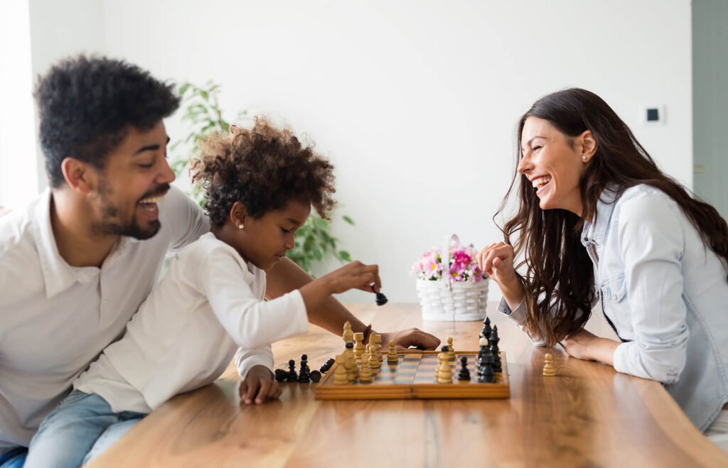 A biracial family sitting at a wooden table smiling & playing chess. Discover how counseling in Katy, TX can help you navigate stress, anxiety, trauma & more. Get started with WHCC today.