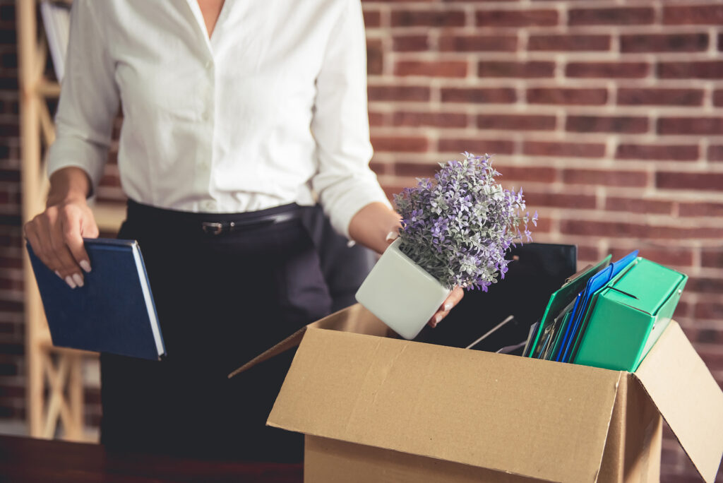A businesswoman packing away purple flowers & a book into a box. Representing life transitions & how taking a new job can bring up mixed feelings. Learn how counseling in Katy, TX can help.