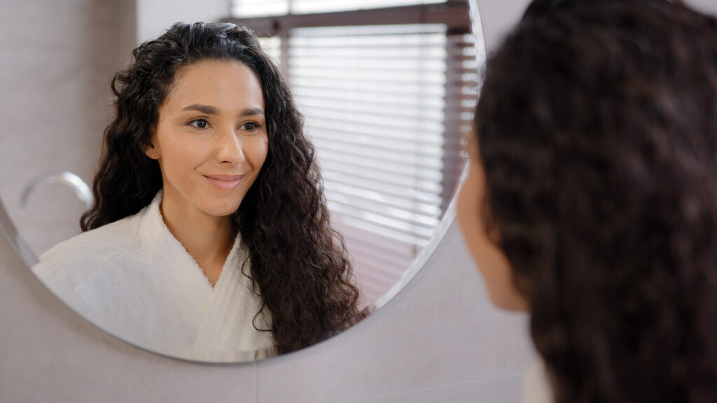 A woman with brown curly hair smiling gently in the mirror. Finding a therapist in Katy, TX is important for your tailored healing journey. Reach out today for counseling.
