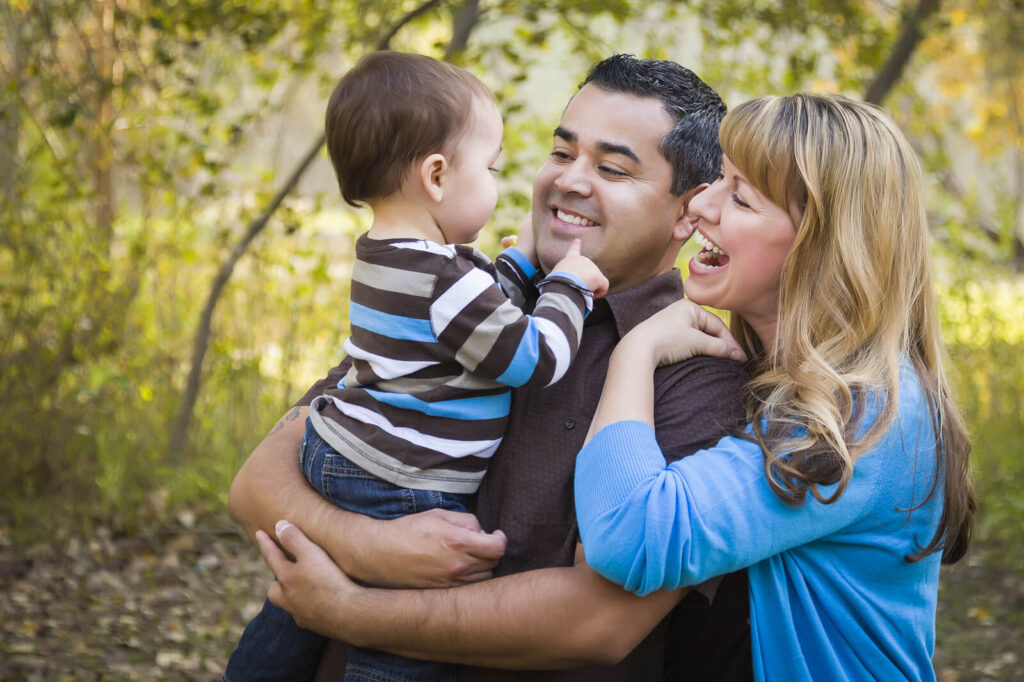 A happy biracial couple holding their baby outside near trees. Adult therapy in Katy, TX helps individuals navigate attachment style challenges & issues. Get started today!