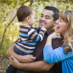 A happy biracial couple holding their baby outside near trees. Adult therapy in Katy, TX helps individuals navigate attachment style challenges & issues. Get started today!