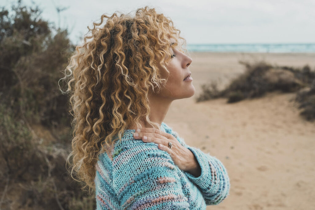 A woman with curly hair taking a deep breath on the beach. Complex PTSD can be treated with the right support. Reach out today to get started with a trauma therapist in Katy, TX.